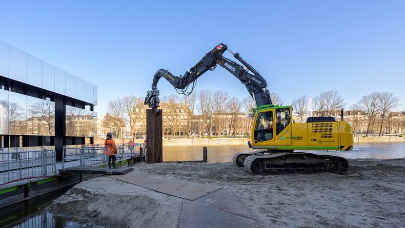 Damwand van het werkgebied half in de grond, met links de spiegelende bouwkeet ern rechts het Eerste Kamergebouw
