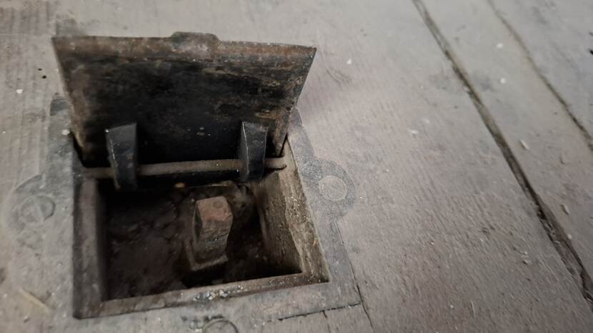 Luikje in houten vloer met daarin een metalen bedieningsscharnier