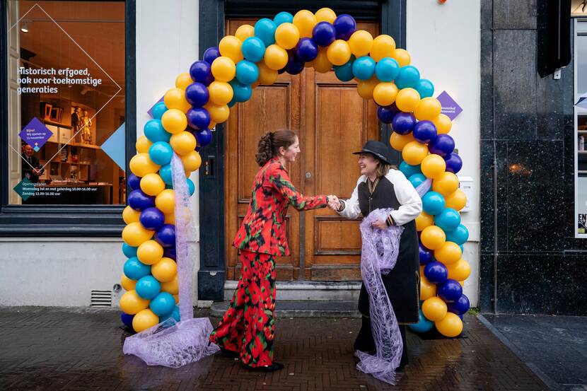 Yvonne van der Brugge-Wolring en Saskia Bruines feliciteren elkaar na het losmaken van een grote paarse strik voor de deur van het Informatiecentrum Binnenhof Renovatie